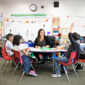 Photograph of a female elementary teacher sitting at a table with 4 young students, one of whom has their hand raised.