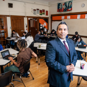 Photograph of a male teacher wearing a suit standing in a classroom with students sitting at tables working on assignments.